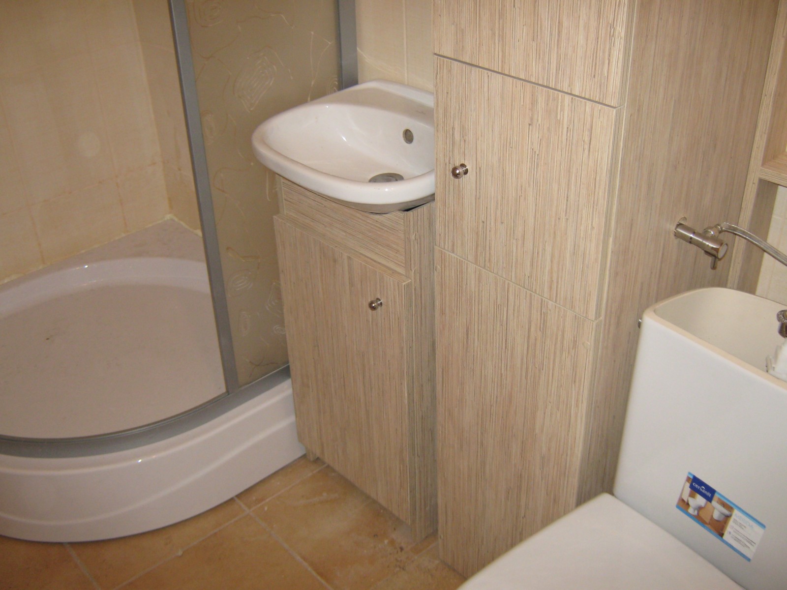 Łazienka z kabiną prysznicową i umywalką z szafką oraz wysoką szafką, wykończone w okleinie imitującej jasne drewno z wyraźnym usłojeniem.